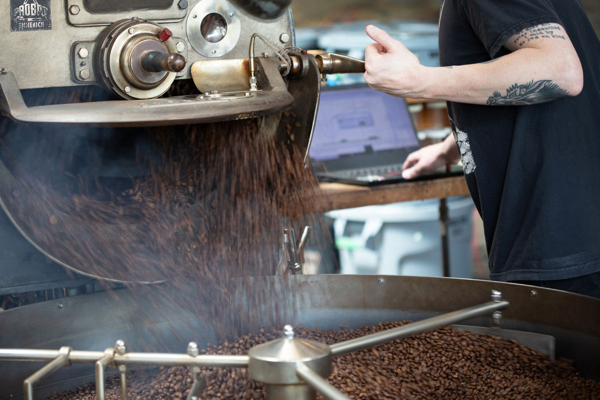 Roasted coffee beans falling from a large metal roasting machine into a tray below, with a person in a black shirt standing at a laptop in the background.
