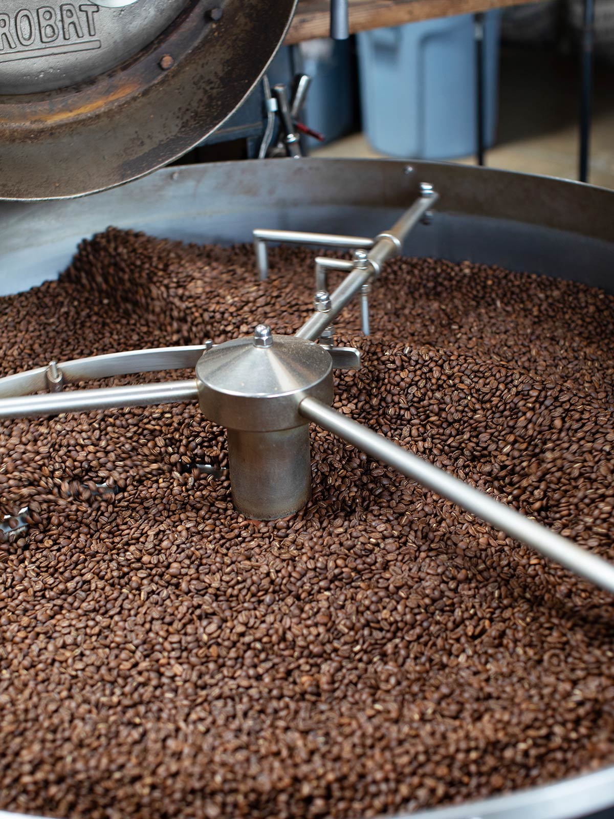 Coffee beans being processed in a large metal container with a metallic device on top.