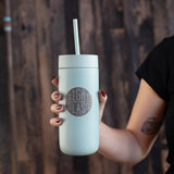 Light blue tumbler with straw held by a person against a wooden background