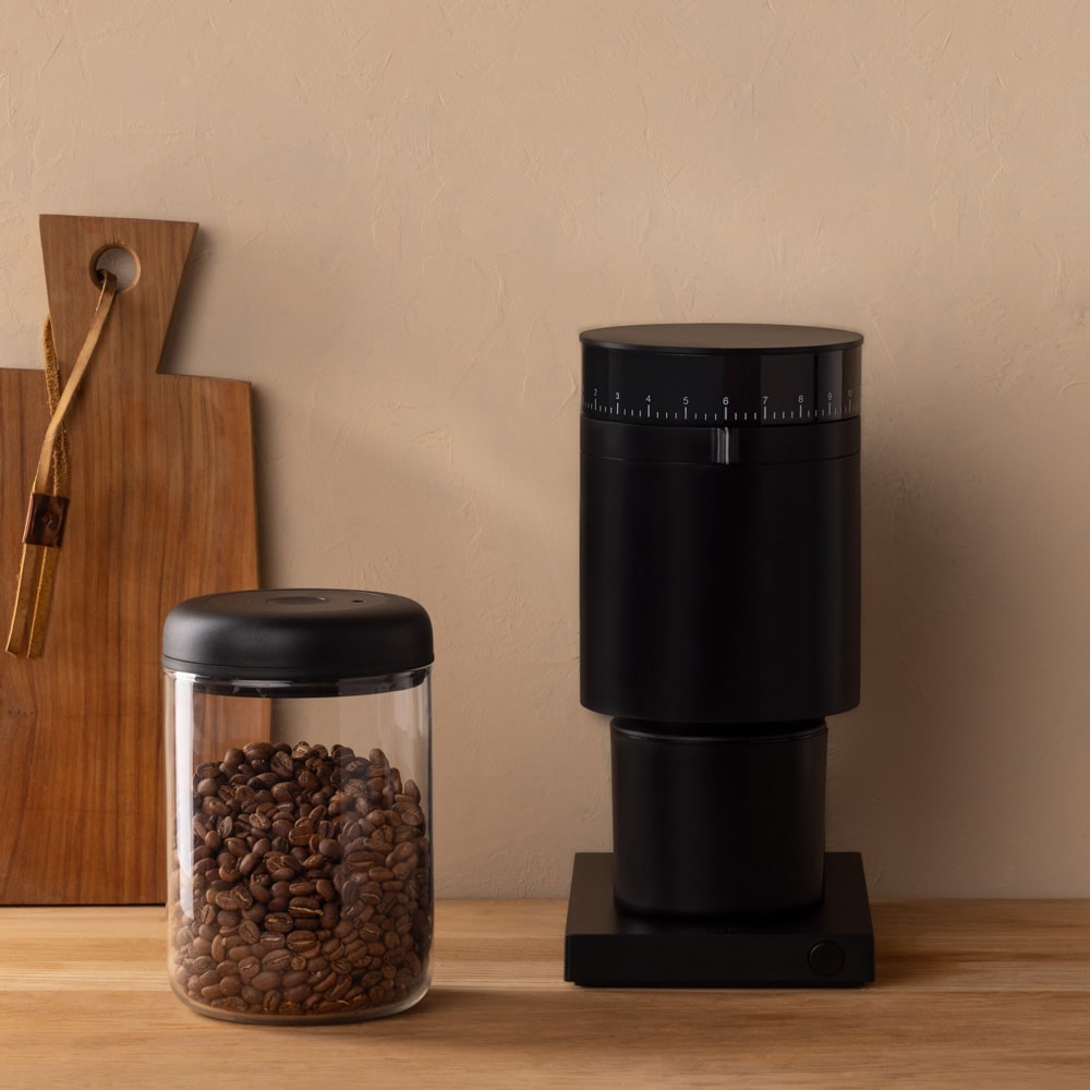 Black coffee grinder and glass container with coffee beans on a wooden surface.